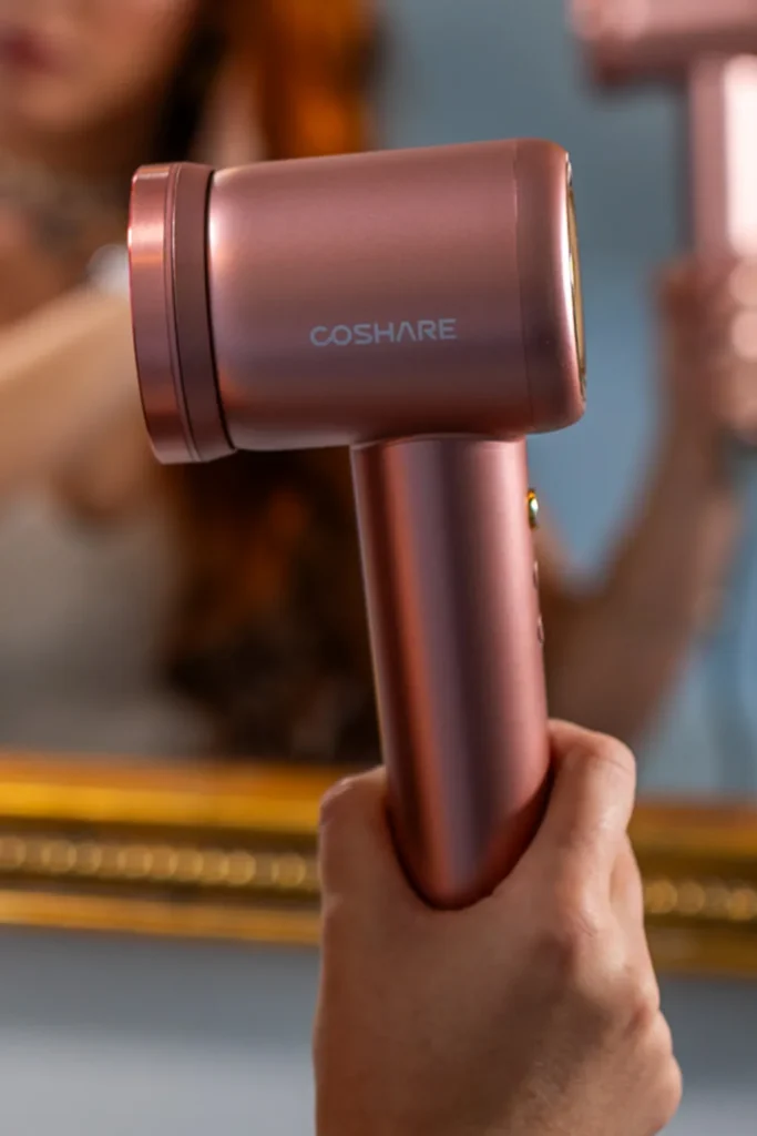 Suszarka do włosów COSHARE SuperFlow1 PRO Rose Gold trzymana w dłoni, w tle lustro